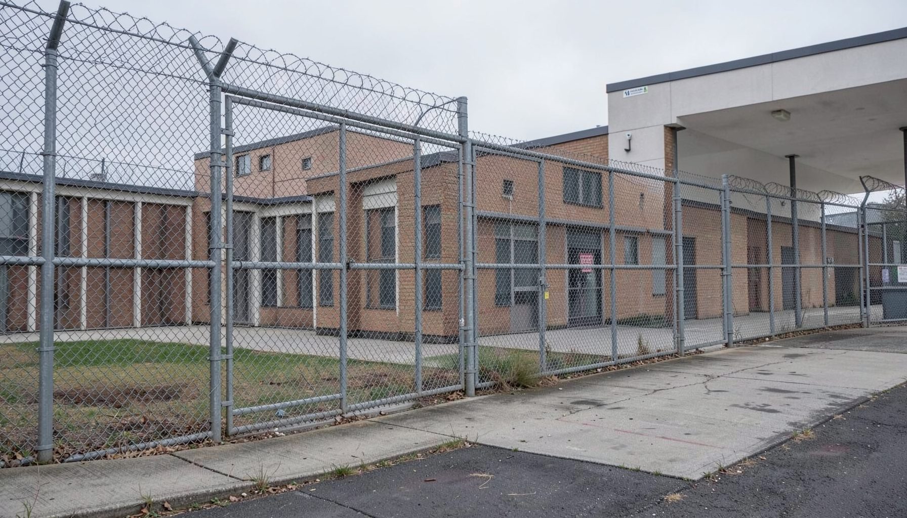 A correctional facility exterior with fencing, institutional architecture, sober mood.