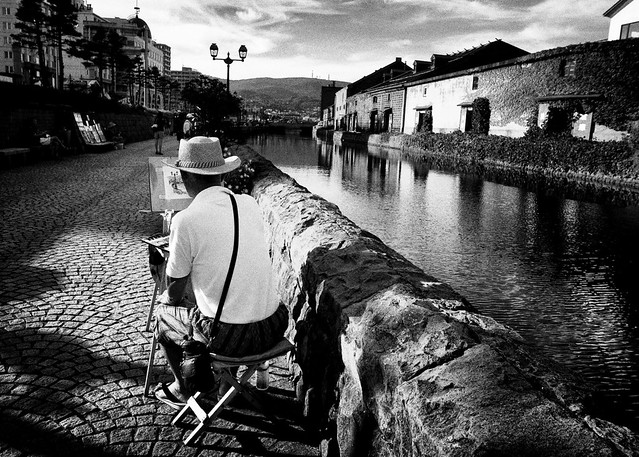 Person in a hat painting beside a canal lined with old brick buildings, black and white, calm reflective mood.